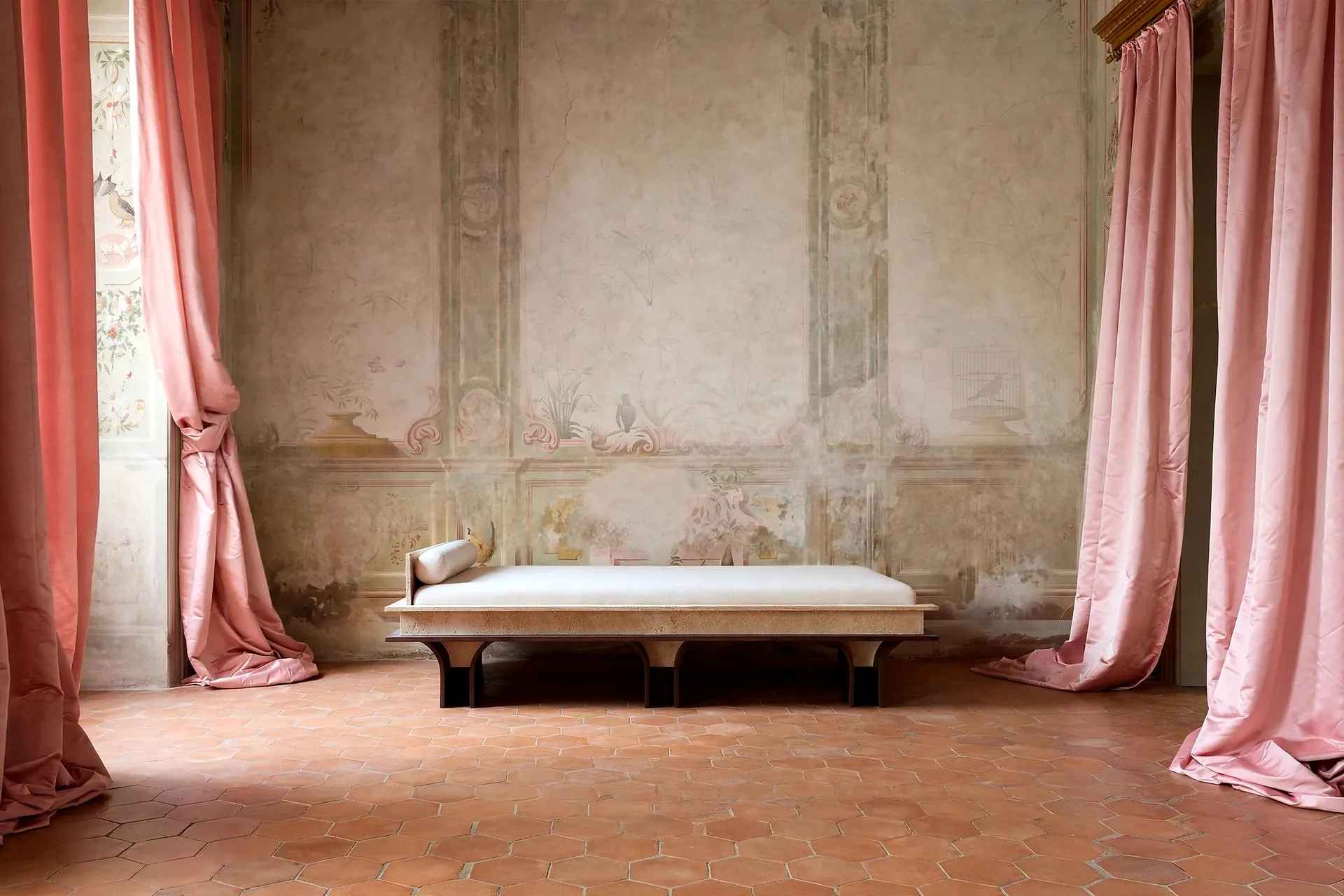 Arcus daybed in stone and wood in a room with faded frescoed walls, pink silk curtains and a terracotta hexagonal floor